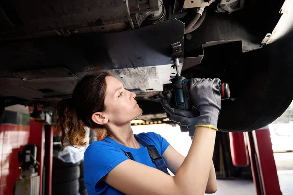 Woman auto mechanic works under a car raised on a special lift, the master uses a pneumatic impact wrench