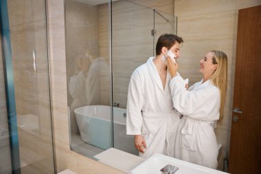 Mutlu bir eş kocalarının yanaklarına tıraş köpüğü sürüyor. Çift sabah banyoda rutin şeyler yapıyor.