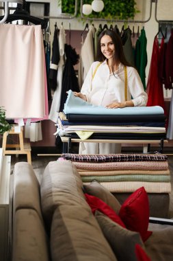 Kadın tasarımcı kumaşlarla dolu bir masada duruyor. Stüdyoda çok çeşitli malzemeler var.