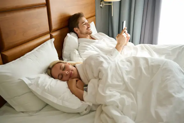 Guy Mobile Phone Resting Large Bed Cute Blonde Dozing Nearby — Stock Photo, Image