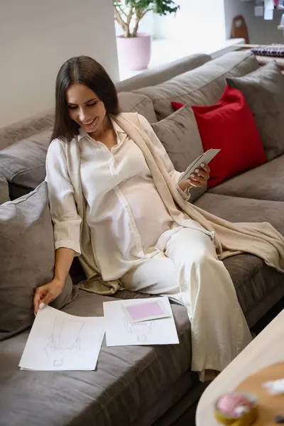 Pregnant woman studies clothing sketches in a design studio, she sits on a comfortable sofa