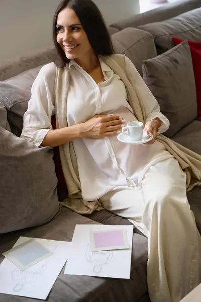 Smiling pregnant female sits on the sofa with a cup of coffee, clothing sketches lie next to her