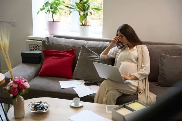 Pregnant woman with poor health is working on a laptop, she is sitting on a large sofa