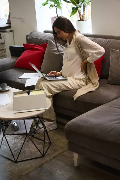 Designer with poor health is working on a laptop, she is sitting on a large sofa