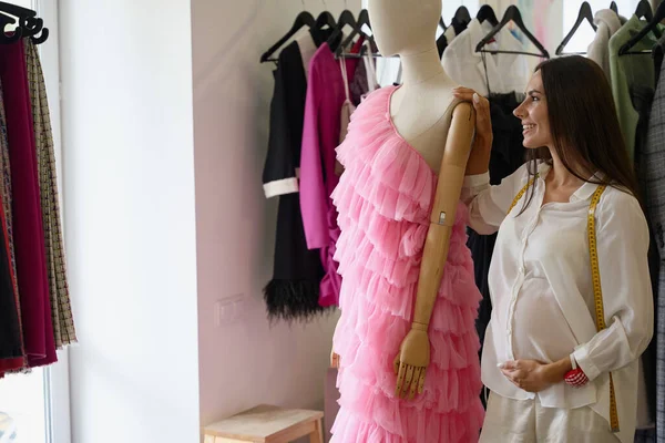 Pregnant woman stands by a mannequin in a pink evening dress, she has sewing supplies