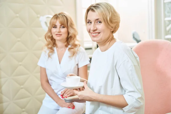 笑顔の大人の白人女性 お茶やコーヒーと美容院でカメラを見ているぼやけた美容師 — ストック写真