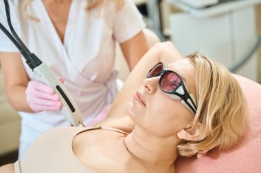Kısmen kadın güzellik uzmanı, IPL fotorejuvenation cihazıyla lazer epilasyon yapıyor. Bulanık güzellik salonunda gözlük takan yetişkin beyaz kadın koltukaltı. Vücut bakımı kavramı