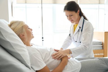 Konsantre olmuş beyaz kadın doktor doğum kliniğinde Avrupalı kadının hamile karnını kontrol ediyor.