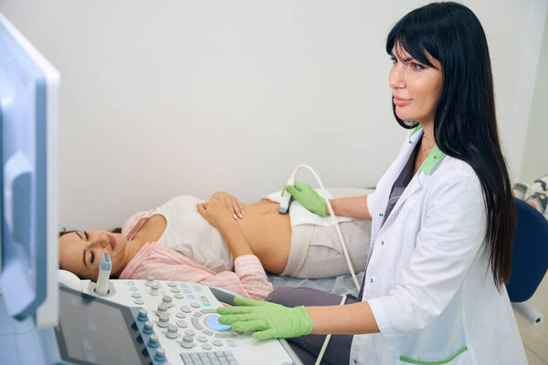 Adult european female gynecologist doing ultrasound examination to young caucasian pregnant woman in clinic. Concept of pregnancy examination