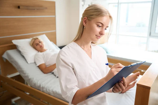 産科病院の医療ベッドに横たわるぼやけた妊婦の背景を持つクリップボードに焦点を当てた白人女性看護師のフロントビュー — ストック写真