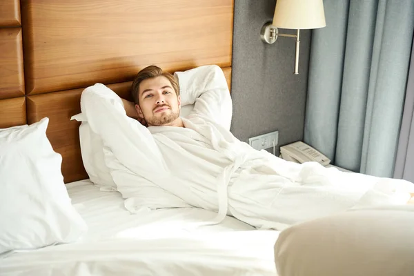 Hotel guest in a bathrobe rests on a large bed, the man has a small beard