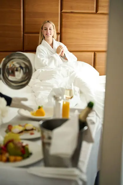 Hotel worker serves breakfast to a happy lady in her room, female in a cozy terry robe