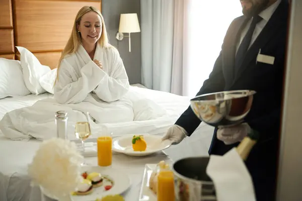 Hotel worker serves breakfast to a young woman in her room, a lady in a cozy terry robe