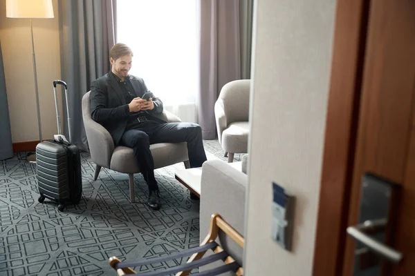 Smiling man sits with a mobile phone in a hotel room, next to a travel suitcase
