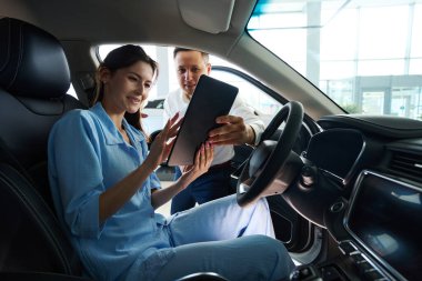 Araba galerisi müdürü yeni bir araba için belgeleri müşteriye sunar, bayan sürücü koltuğunda oturur.