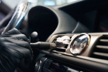 Koruyucu eldiven giyen usta arabanın içindeki havalandırmayı özel bir fırçayla temizliyor, gösterge panelinde bir saat var.