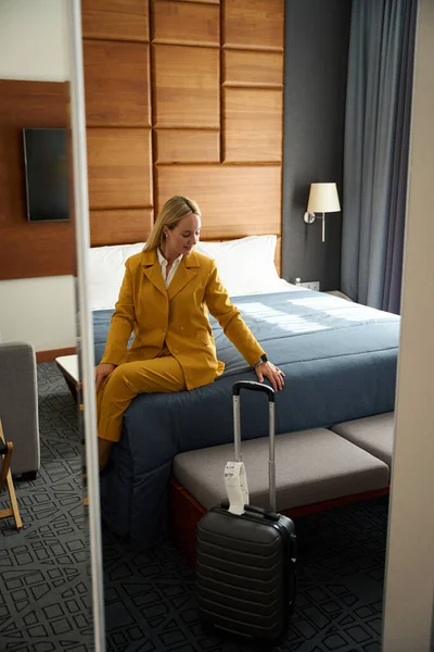 Young female in a yellow travel suit sits on large bed in a hotel room, next to a travel suitcase