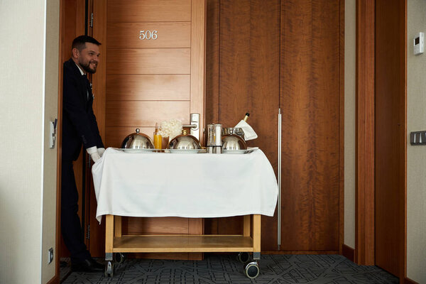 Smiling waiter delivered food to the room on a serving table, the food is covered with cloche lids