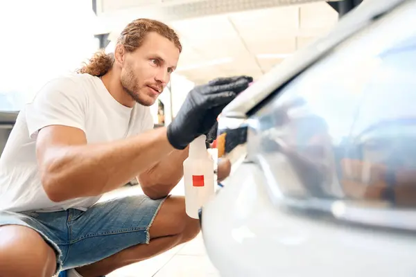 Long-haired male uses special clay in car detailing, a spray bottle is used in the work