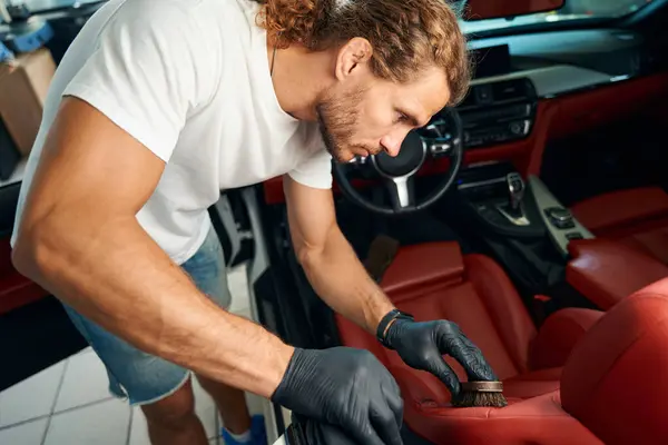 Long-haired guy cleans a leather drivers seat with a brush, he is doing detailing of the car interior