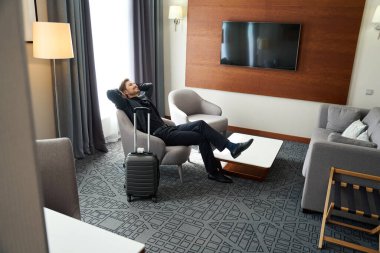 Mutlu adam, kollarını bir otel odasında bir seyahat çantasının yanına uzatarak oturur.