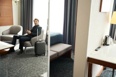 Seyahat kıyafeti giymiş bir adam koltukta cep telefonuyla oturuyor, seyahat çantasının yanında.