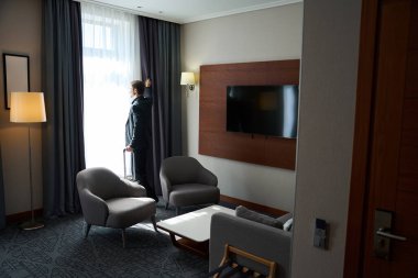 Seyahat elbiseli bir adam bir otel odasının penceresinin yanında durur, oda modern bir minimalist tasarıma sahiptir.