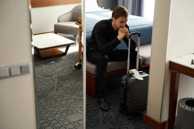 Seyahat kıyafetleri içinde üzgün bir adam otel odasında bir yatağa oturur, oda modern bir minimalist tasarıma sahiptir.