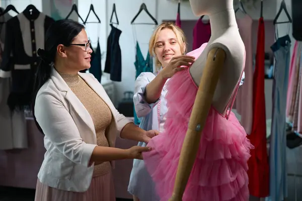Two women stylists model an evening dress on a mannequin, there are many clothing samples in the room