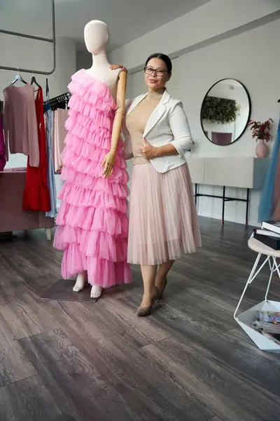 Beautiful woman stands by a mannequin in a sewing studio, the mannequin is wearing an evening dress