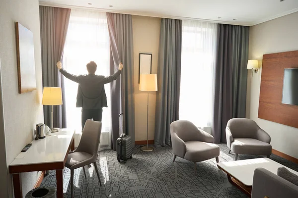 Man in travel clothes opens the curtains in a hotel room, the room has a modern minimalist design