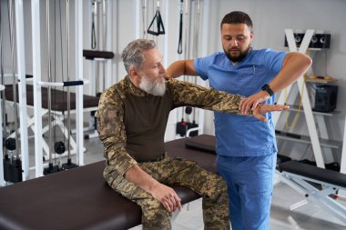 Askeri giysili bir adam hastanede rehabilitasyon uzmanıyla görüşüyor. Adamlar spor salonunda oturuyor.