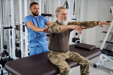 Rehabilitolog askeri giysili bir adamın arkasını inceliyor, hasta masaj masasında oturuyor.