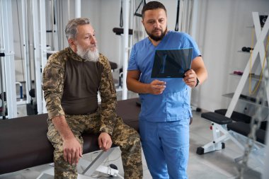 Spor salonunda iki sakallı adam röntgenlere bakıyor, modern ekipmanlar odada.