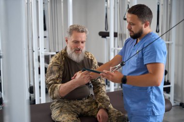 Doktor rehabilitasyon uzmanı askeri bir hastayla spor salonunda çalışıyor. Kamuflaj kıyafeti giymiş bir adamla.