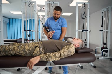Sakallı adam kol kaslarını özel bir simülatöre germek için egzersiz yapar. Rehabilitasyon uzmanı ona yardım eder.