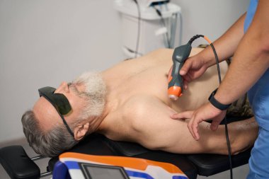 Tıbbi rehabilitasyon uzmanı askeri bir hastayla çalışıyor. Güvenlik gözlüğü takan bir erkek kanepede yatıyor.