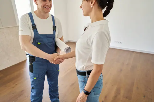 Obscure faces of smiling caucasian male worker with blueprint and woman handshaking and looking at each other in new modern townhouse before repair. Concept of work deal