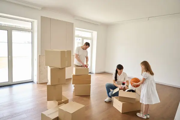 Married couple with a daughter unpacks things in a new house, the girl takes a ball out of the box