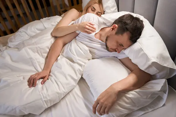 Dozing woman hugs an upset man, the couple sits on a comfortable bed