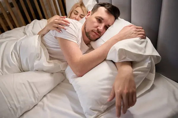Dozing wife hugs her upset husband, the couple sits on a comfortable bed