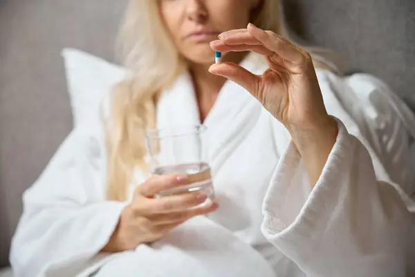 Cropped photo of woman in bathrobe seated in bed holding glass of water and medicine capsule in hands