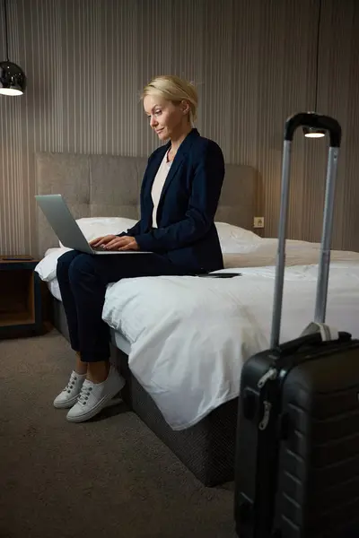 Focused businesswoman in pantsuit seated on bed in hotel room typing on her portable computer