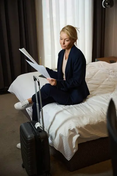 Serious focused businesswoman seated on bed in suite with pencil in hand looking through business documents