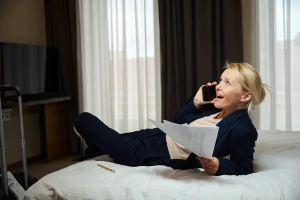 Cheerful business lady with pile of documents in hand lying on bed in hotel room during smartphone conversation