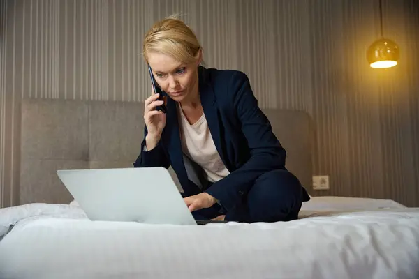 電話会話中にポータブルコンピュータで作業するスイートでベッドに座った集中ビジネス女性 — ストック写真