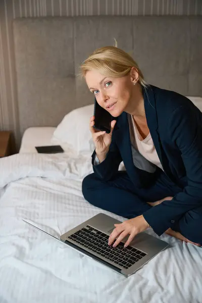 Calm businesswoman sitting on bed in suite while typing on laptop keyboard during phone conversation