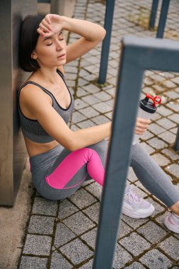 Metal yapıların yanında oturan bir kadın, su şişesi tutuyor ve dışarıda sıkı bir eğitimden sonra dinleniyor. Spor konsepti