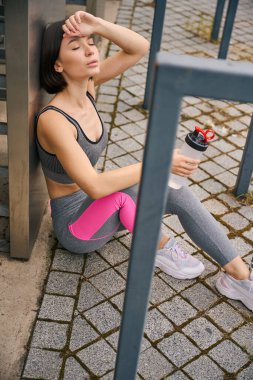 Sportif kadın antrenmandan sonra yorgun, metal yapının yanında oturuyor, elinde bir şişe su tutuyor.