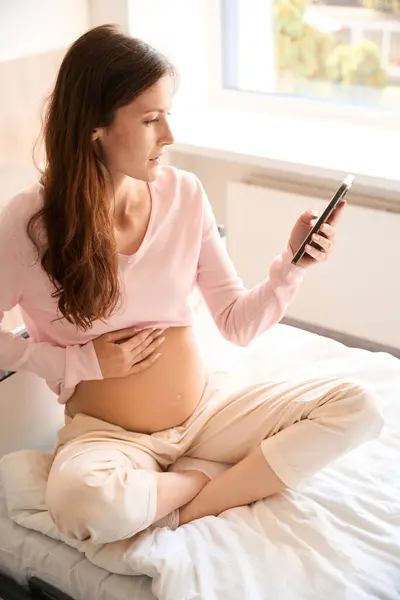 Pregnant woman using app on smartphone for checking interval between contractions on bed in clinic